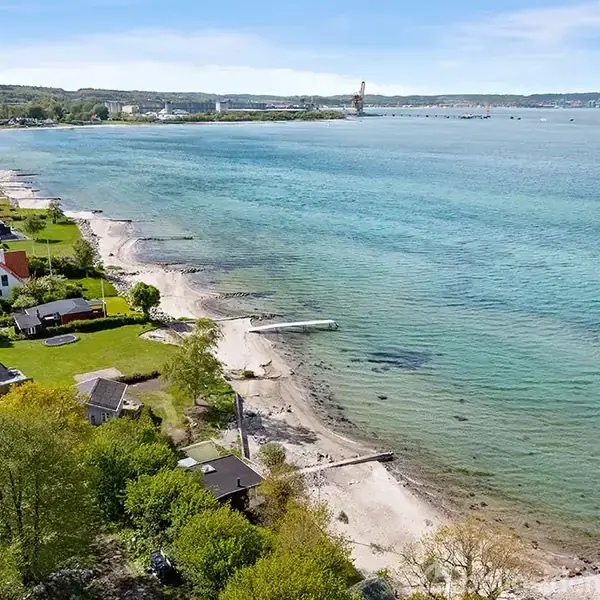 Kystlinje med huse ved vandet, nogle med haver, strækker sig langs strand. Dybt blåt hav med klart vand spreder sig mod horisonten under en lys himmel.