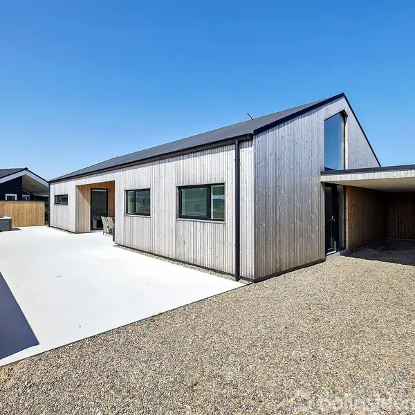 Et moderne hus med træfacade står på en solrig dag. Huset har en bred indkørsel af beton og en carport. Området er roligt med få nærliggende bygninger.