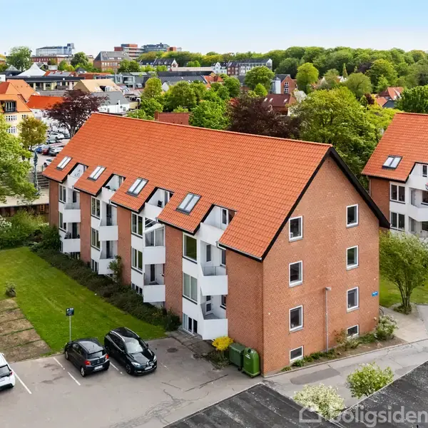Et rødt murstensbyggeri med skråt tag og flere vinduer står ved en parkeringsplads. Flere biler er parkeret foran bygningen, omgivet af andre boliger og træer i et byområde.