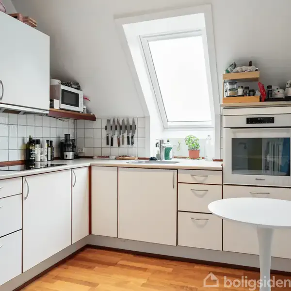 Hvidt køkken med skabe, mikroovn, knivholder og potteplante. En hvid rund bord foran. Ovenlysvindue lader naturligt lys strømme ind. Blå forklæde hænger på væggen til højre.
