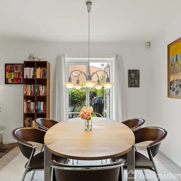 Et rundt spisebord med seks stole står under en hængelampe i en stue med hvide vægge. I nærheden er en bogreol, sofa og et stort farverigt maleri.