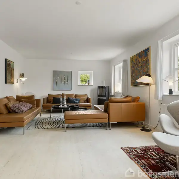 Stue med brune lædersofaer placeret omkring et sofabord, mens en lampe står tændt ved siden af. Omkring ses moderne kunstmalerier, hvide vægge og store vinduer med udsigt til haven.