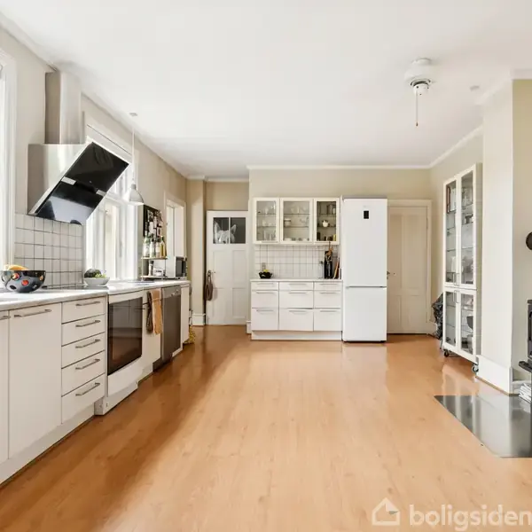 Et lyst køkken med hvidt interiør og trægulv. Køkkenet har store vinduer og moderne apparater, inklusive en ovn og emhætte. I hjørnet står en brændeovn, skaber varme og hygge.