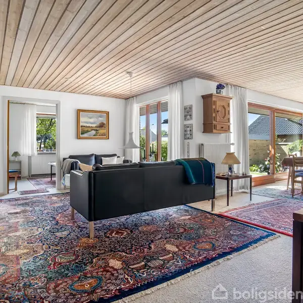 Stue med sorte sofaer placeret på orientalske tæpper. Store vinduer slipper lys ind og afslører udsigt til en terrasse. Træloft og lyse vægge skaber en rummelig atmosfære.