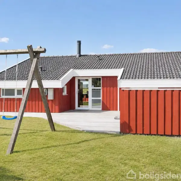 Rødmalet træhus med sort tag står på græsplæne. Ved siden af, gyngestativ med to gynger. Klar blå himmel i baggrunden.