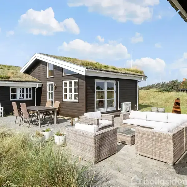 Sort sommerhus med græs på taget står i klitlandskab. Foran huset er der udendørs møbler og terrasseområde med fliser. Himlen er blå med spredte skyer.