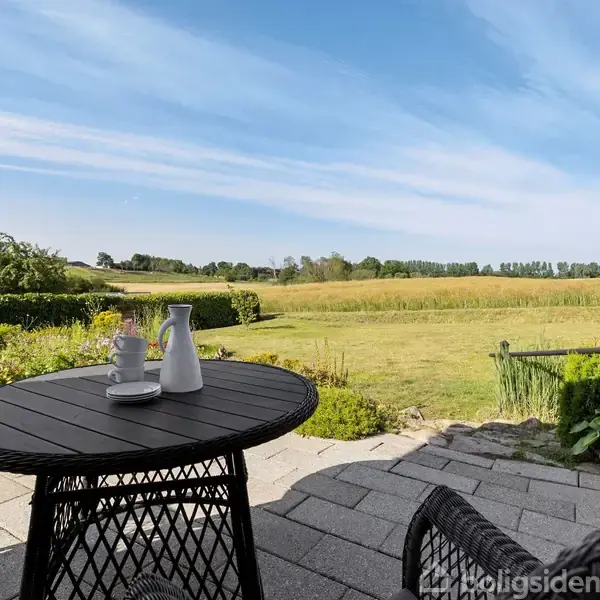 En rund sort havebord står med to kander på en stenbelagt terrasse. Ved siden af står sorte stole. I baggrunden ses en grøn mark og klar blå himmel.