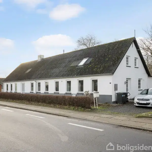 Hvidt, langstrakt hus med saddeltag står langs en vej. Ved siden af huset er en hvid bil parkeret på en grusbelagt parkeringsplads. Der er hække foran huset.