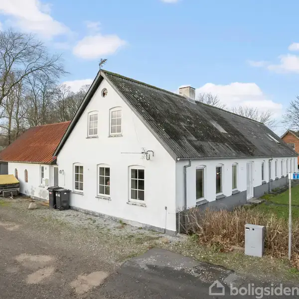 Hvid bygning med rødt tag står ved siden af en vejskilt med teksten "Præstegårdsvej". Omkring bygningen er en grusvej og nogle træer, der danner en landlig omgivelser.