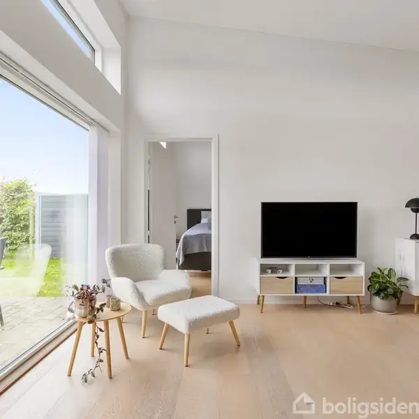 En lys stue med lyse træmøbler og en fladskærms-tv på et hvidt skab. Store glasdøre fører ud til en terrasse med grøn have. Et værelse ses i baggrunden.