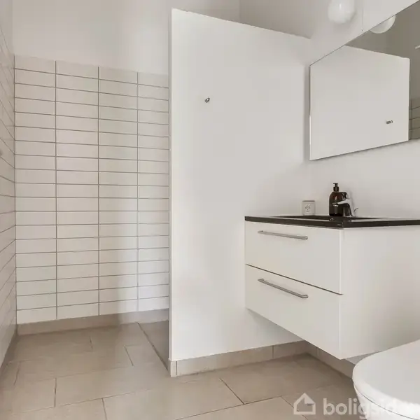 Badeværelse med hvidt flisegulv og vægge. Skabet under vasken har to skuffer. Øverst til højre er hylder med forskellige flasker. En moderne, minimalistisk indretning.