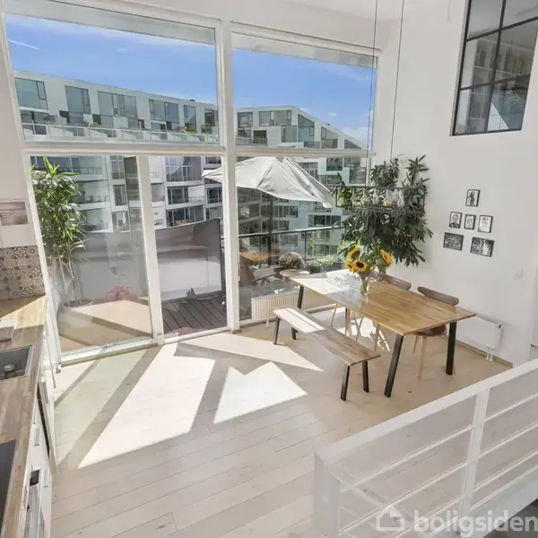 Lyst køkken-alrum med træbord og solhattekande på. Store vinduer lader sollys strømme ind. Udsigt til moderne lejlighedskompleks. Dør fører til soveværelse. Hvidt interiør med grønne planter.