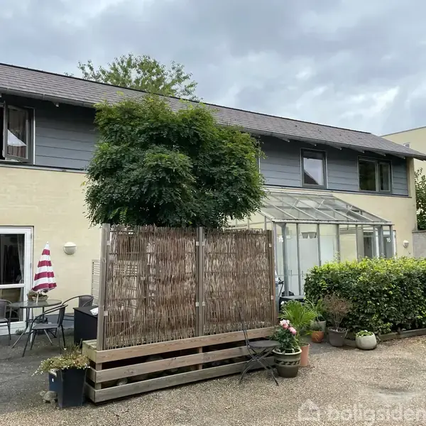 Bygning med to etager, omkranset af have med planter og havemøbler. En glasveranda er tilknyttet nederste etage. Overdækket himmel indikerer overskyet vejr.