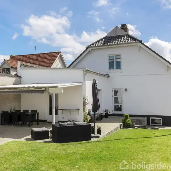 Hvidt hus med sort tag står i en have med hække. En terrasse med møbler og parasol er delvist overdækket, mens en grill står i nærheden. Solrig dagslys.