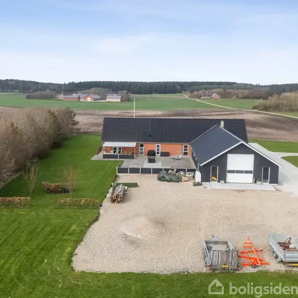 Et rødt murstenshus stands stille med en tilstødende garage på en grusplads, omgivet af grønne marker og en række træer på den ene side. En rød traktor står parkeret.