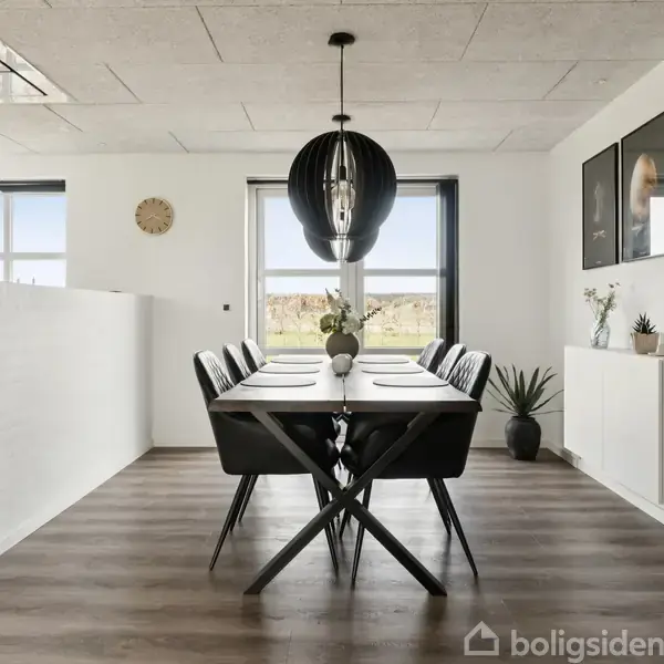 Spisebord med sorte stole står i en lys spisestue. Bordet er dækket og omgivet af moderne indretning med planter og vægkunst. En stor pendellampe hænger over bordet.