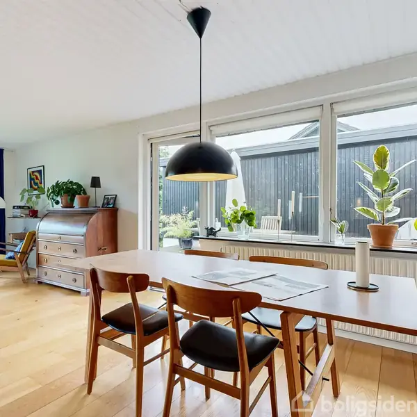 Spisebord med fire stole står ved stort vindue, hvorfra der ses en træterrasse. En sort lampe hænger over bordet, og planter pryder rummet.