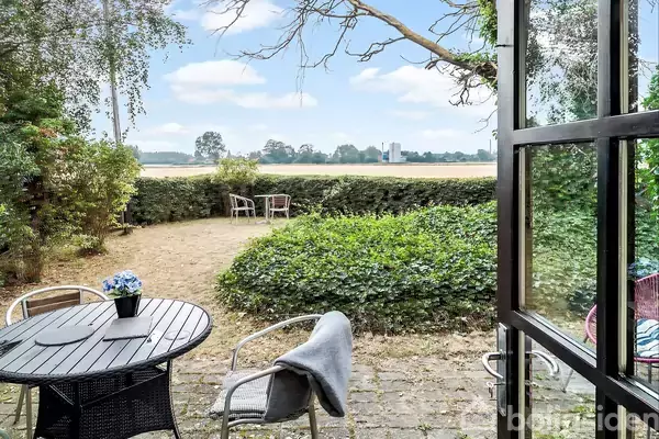 Et rundt bord med en blå blomstervase står på en terrasse. To stole har et tæppe over ryggen. I baggrunden ses en græsplæne og marker under en blå himmel.