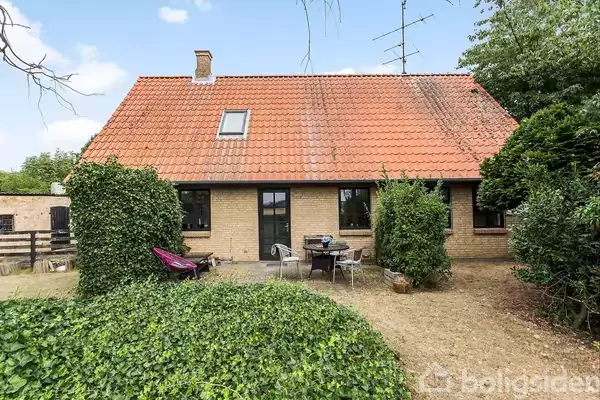 Et hus med rødbrunt tegltag har en terrasse foran, hvor der står et bord og stole. Haven omkring er grøn og tilgroet med buske og træer.