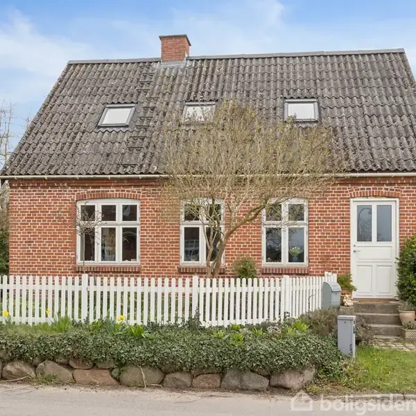 Rødt murstenshus med gråt tegltag, hvide sprossevinduer og hvid indgangsdør. Foran står et hvidt stakit og nogle trappetrin, omgivet af buske og blomster under en blå himmel.