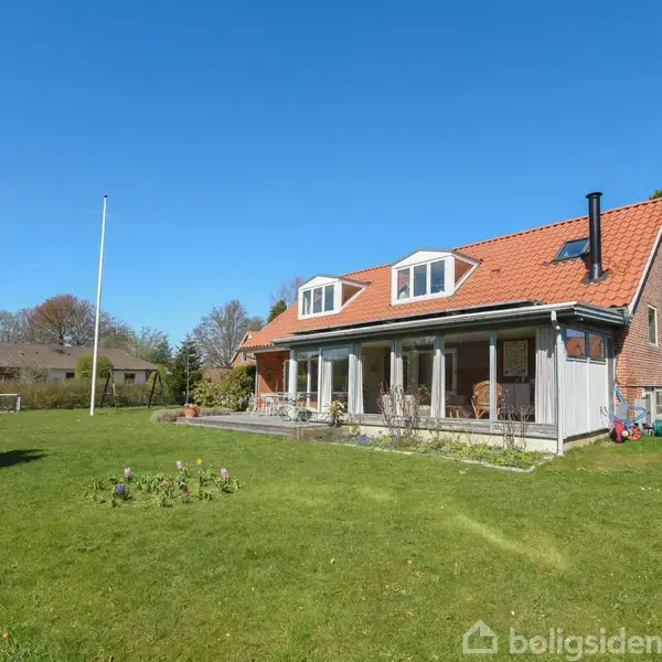 Hus med rødt tag og kviste står stille på grøn græsplæne omgivet af træer og klar blå himmel. Terrasse med glasvægge ses på siden af huset.