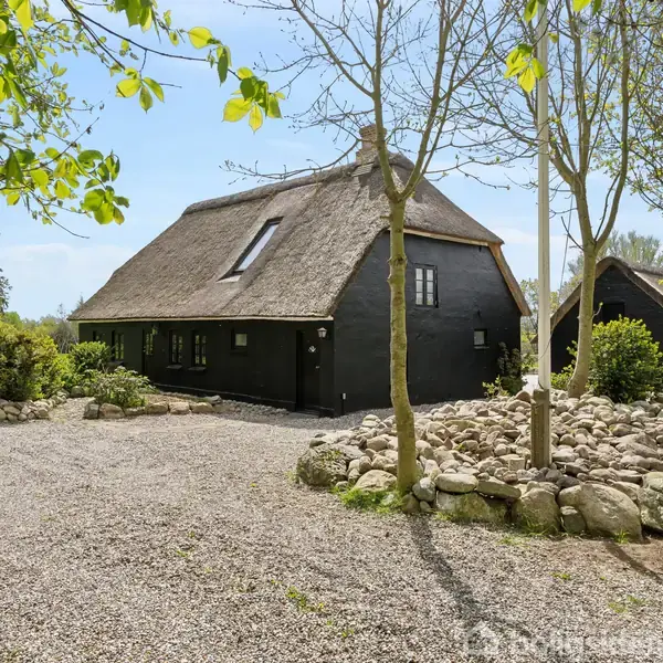 Stråtækt hus står stille i en solrig, frodig have omgivet af træer og en grussti. Store sten danner en dekorativ kant ved siden af huset.