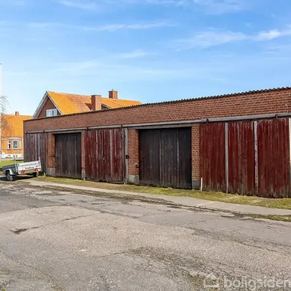 En lang, lav bygning med røde træporte står langs en vej. En gammel bil holder parkeret foran bygningen, omgivet af huse med røde tage på venstre side.