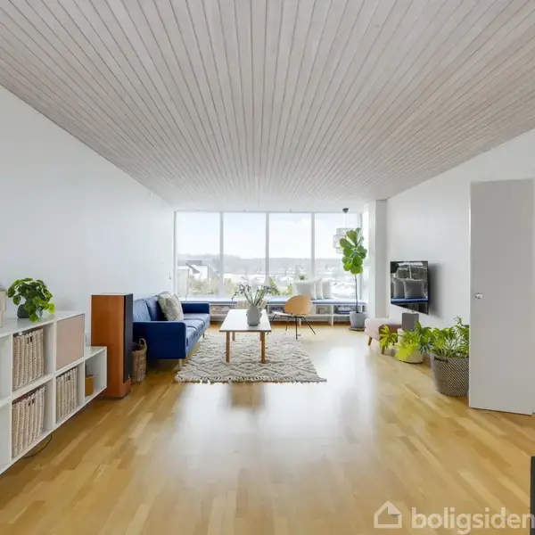 En stue med trægulv og hvide vægge indeholder en sofa, sofabord og potteplanter. Store vinduer giver udsigt over byen. En åben dør fører til et tilstødende rum.