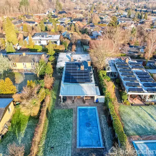 Hus med solpaneler har en udendørs swimmingpool i baghaven. Omgivet af andre villaer i et boligkvarter med træer og græsarealer.