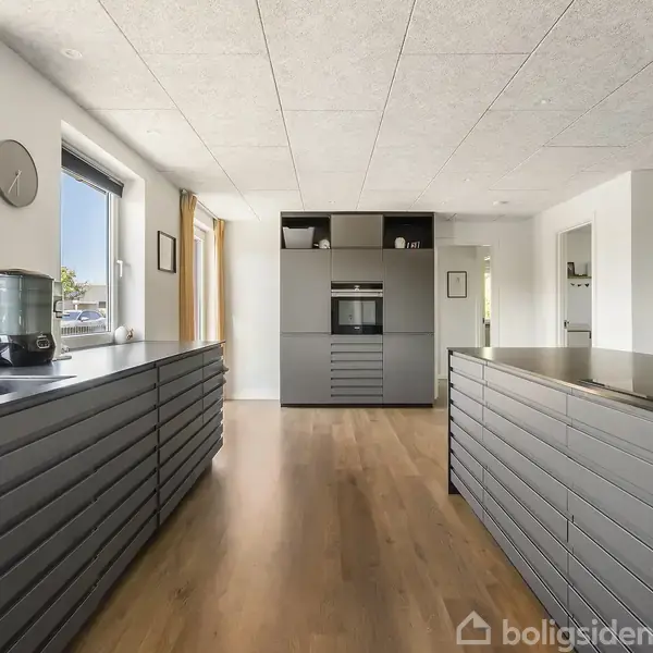 Moderne køkken med lange, grå skabe på begge sider, centralt placeret ovn og åbne reoler. Store vinduer til venstre giver naturligt lys. Trægulv og hvide vægge skaber et lyst rum.