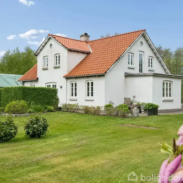 Hus med rød teglstenstag og hvid facade; omgivet af grøn græsplæne og buske; under blå himmel med skyer; blomst i forgrunden til højre.