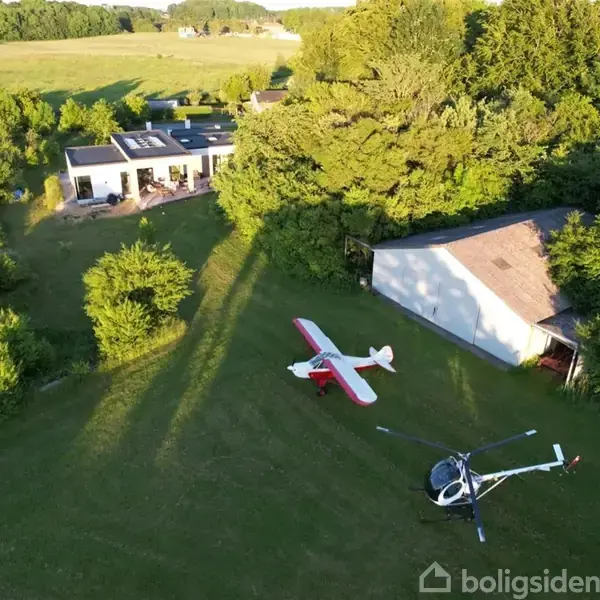 Et lille fly og en helikopter står parkeret på en grøn græsplæne. I nærheden er der et hvidt hus og en lade omgivet af træer og marker.