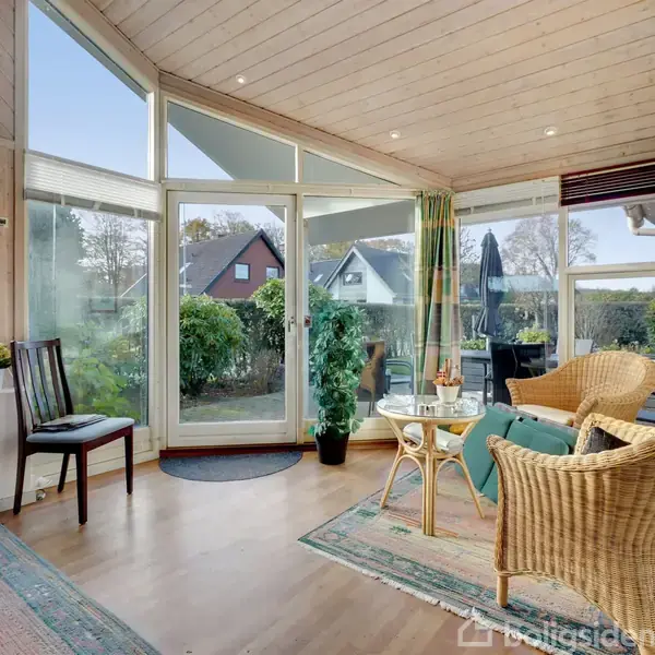 En lyse stue med kurvemøbler og tæpper placeret på trægulv. Store vinduer og en terrassedør giver udsigt til en have med buske og huse i baggrunden. Ingen tekst til stede.