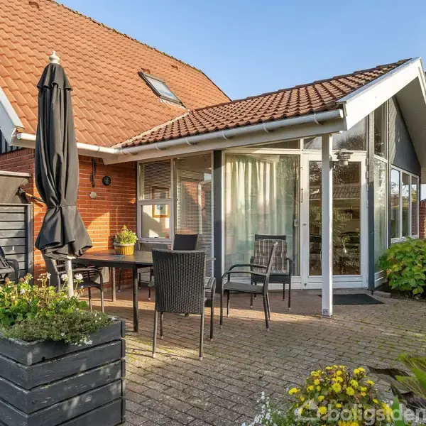 Hus med rødbrune tagsten, stor glasveranda og tilhørende terrasse udstyret med sorte havemøbler og parasol. Omgivet af grønne planter og blomster i plantekasser.