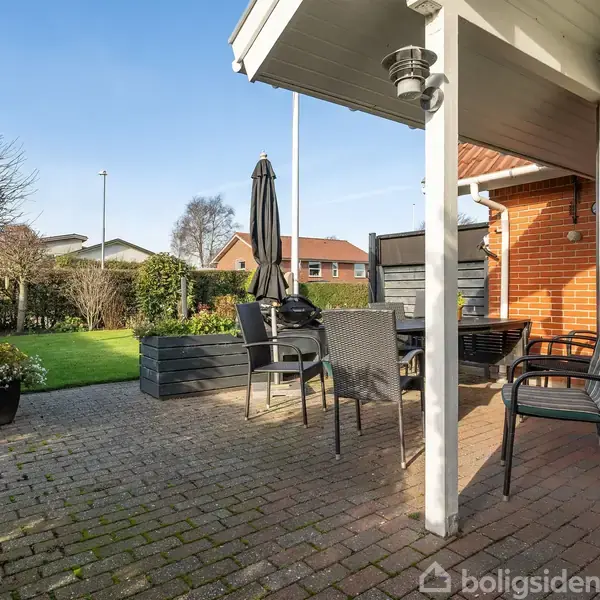 En møbleret terrasse med sorte stole og et bord står foran et murstenshus. En parasol er placeret ved bordet. Græsplæne og buske omkranser området.