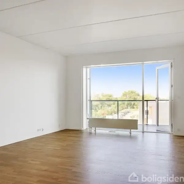 Et tomt rum med trægulve og hvide vægge, har en åbnet altandør, der fører til en balkon med udsigt over et grønt område.