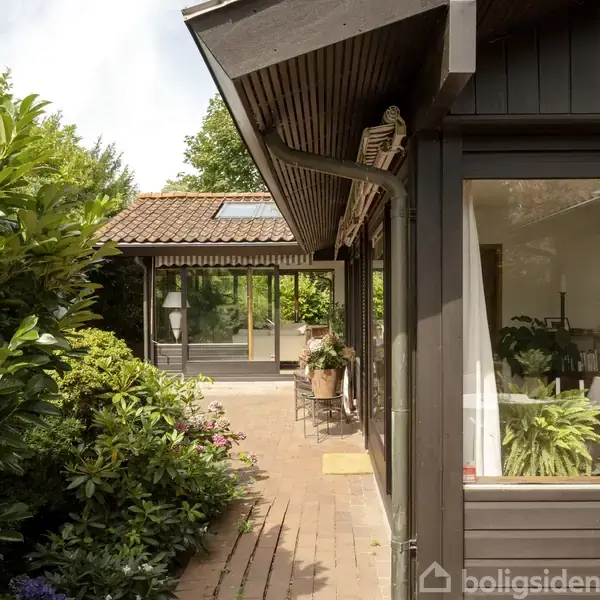 En udestue med store vinduer står i en frodig have, omkranset af grønne buske. Et borde og planter ved siden af et mørkt træhus med skydevinduer.