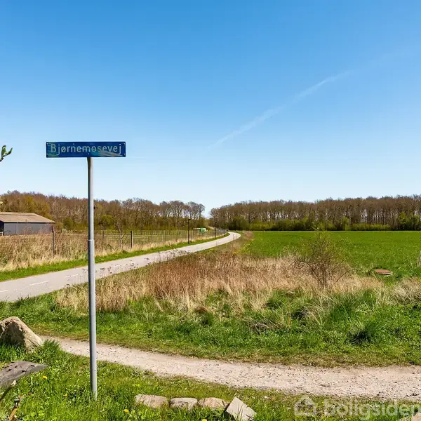 Vejskilte viser "Bjerremosevej" til venstre og bolignummer "20-27" til højre. Omkring ses en landevej, omgivet af marker og træer under en klar blå himmel.