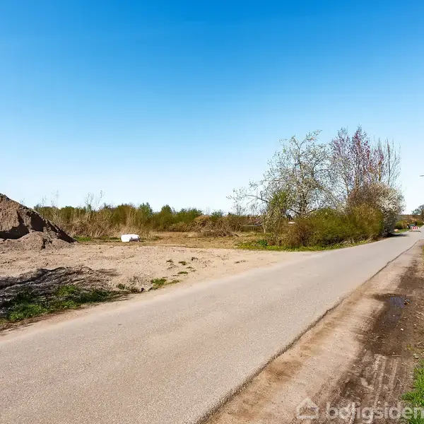En asfaltvej løber gennem et åbent område med jord og grusstabler til venstre, omgivet af træer og buske under en klar blå himmel.