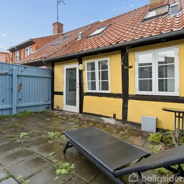 En liggestol står på en flisebelagt terrasse foran et charmerende, gulmalet bindingsværkshus med rødt tegltag. En blåmalet træhegn omkranser gårdhaven, og der vokser små planter mellem fliserne.