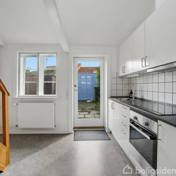 Et lyst køkken med hvide skabe. En trappe i træ fører opad. Gennem en glasdør ses en udendørs have med fliser og en blå skur. Vinduet slipper lys ind.