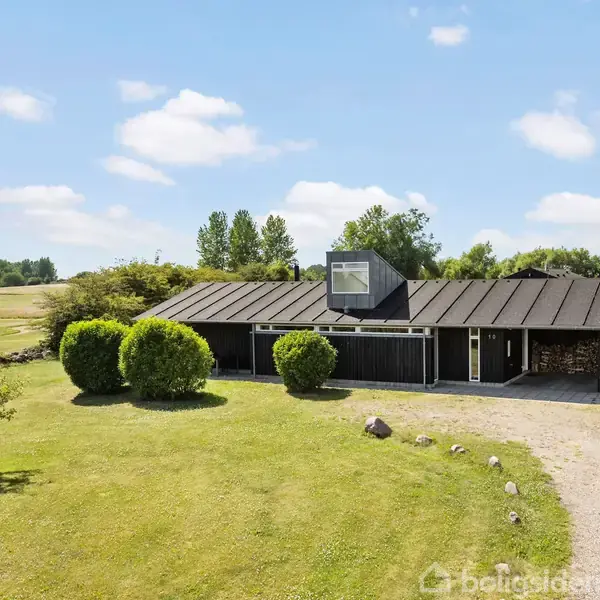 En sort-hus står stille på en velplejet græsplæne med en grusvej foran. Omkring huset er grønne buske og træer under en klar blå himmel.