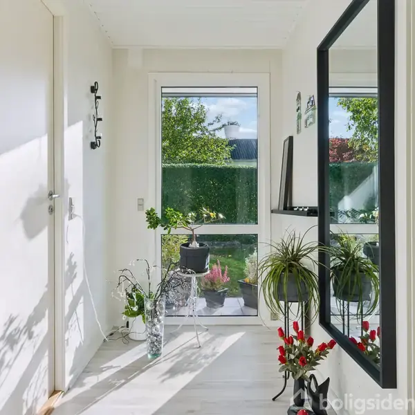 En lys gang med planter placeret på gulvet og et bord, står ved en glasdør. En stor vægspejl hænger til højre, og udenfor ses en grøn have.