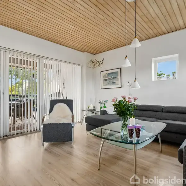 Stue med gråt hjørnesofaarrangement, glas-sofabord med blomster, og en stol med tæppe. Store vinduer med vertikale persienner; træloft og lys trægulv skaber en lys atmosfære.