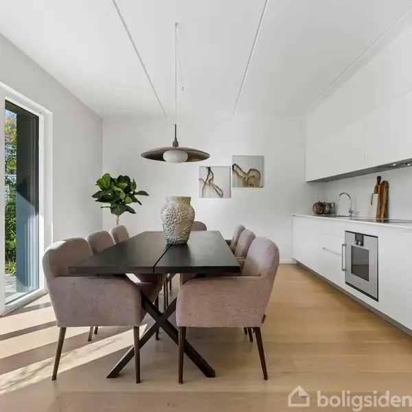 En spisebord med seks stole står i en moderne, lys køkken-alrum med hvide skabe. En stor vase er placeret på bordet. Store glasdøre åbner ud til terrassen.
