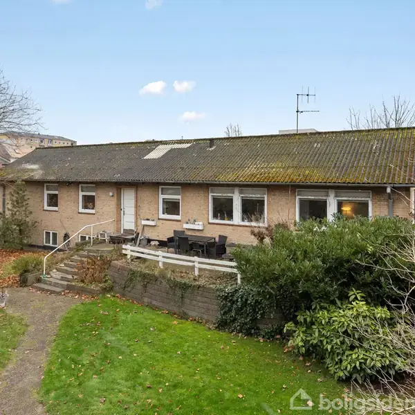 En stor villa står stille i en baghave med grønne græsplæner og buske. Huset har en lang terrasse med havemøbler langs den ene side.