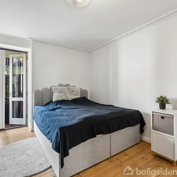 Soveværelse med en dobbeltseng dækket med en mørkeblå dyne. Ved siden af står et natbord med en potteplante. En dør fører til en balkon, og gulvet er af træ.