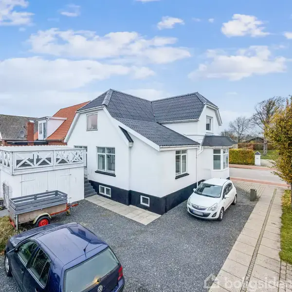Hvidt hus med sort tag står i villakvarter. To biler og en trailer holder parkeret på grusareal. Røde tage og tårn ses i baggrunden, omgivet af fortove og grønne områder.