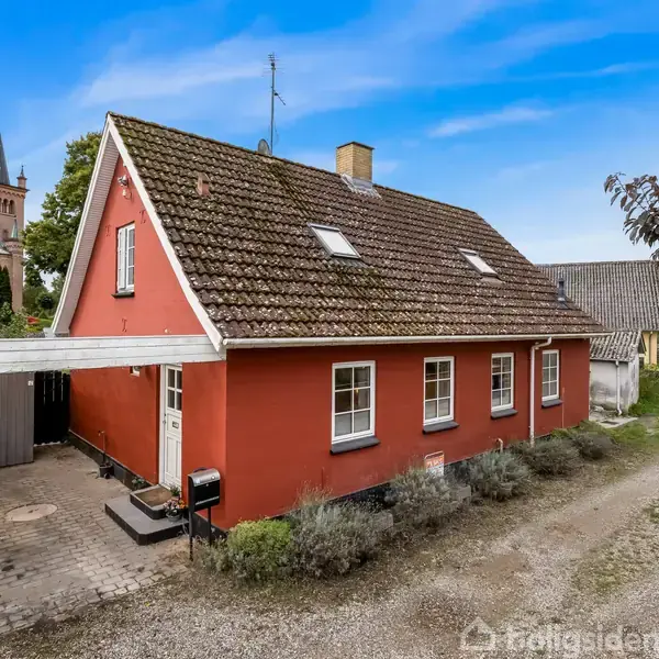 Rødt hus med skråt tegltag og flere vinduer, beliggende ved en smal grusvej. Der er en carport ved siden af huset. I baggrunden ses en kirke omgivet af træer.