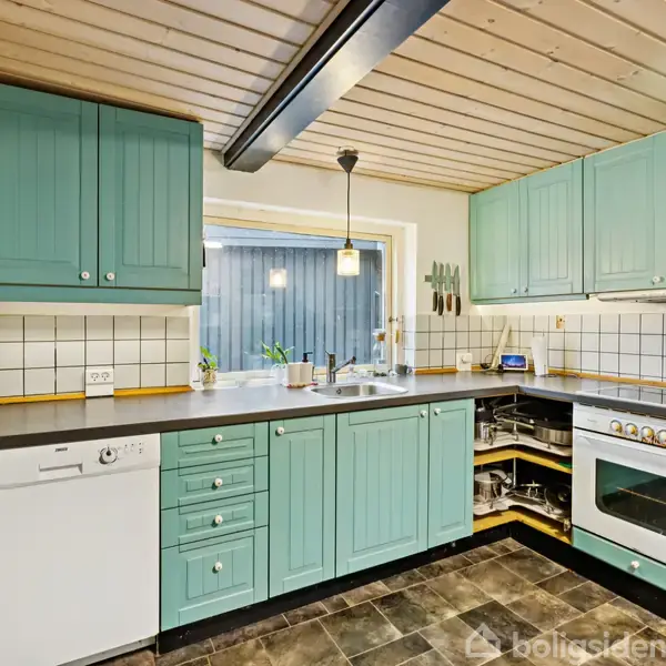 Grønt køkken med skabe og skuffer, udfører funktioner i et hyggeligt rum med sort bordplade, hvid opvaskemaskine, komfur, hvide fliser på væggen og lysentræ loft.